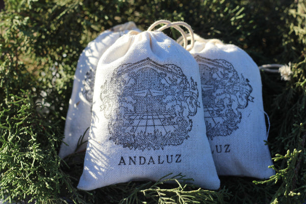 Andaluz Skincare Lavender Sachets made with hand harvested organic Spanish spike lavender . The sachets are off white, made of cotton and have a draw string. This is a photo of them nestled in fir tree branches.